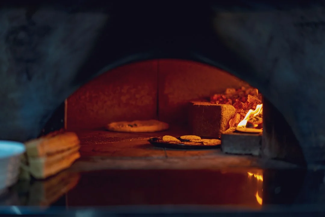 A ceramic artist loading bisqueware onto kiln shelves inside a bright studio, with an L&L electric kiln glowing orange through its peephole in the background